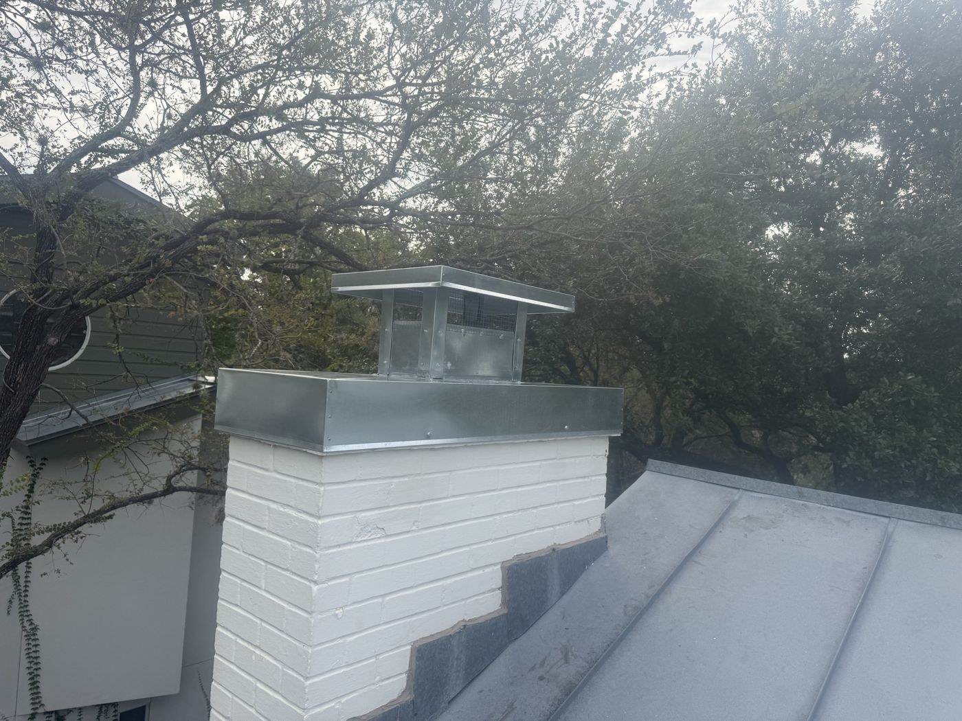 Galvanized chimney cap with mesh on a painted white brick chimney beside a metal roof