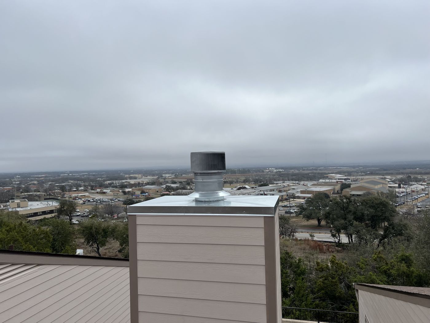 Round galvanized chimney cap on a tan chimney chase with a hillside view